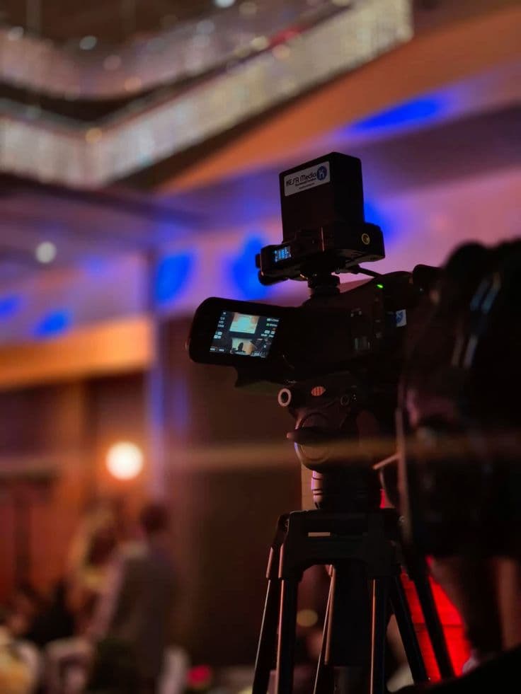 Video camera on a tripod filming an event in a hall lit blue and pink